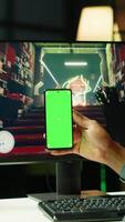 Vertical Man texting friends on green screen phone while playing first person shooter videogame at home. Gamer looking at mockup smartphone display, enjoying online discussion with mates video