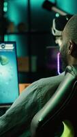 Vertical African american player in neon illuminated apartment paying close attention to computer screen during videogame competitive match. Person competing in internet esports tournament, camera A video