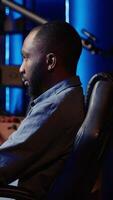 Vertical Portrait of smiling man playing FPS videogame on PC screen in neon illuminated apartment living room. Cheerful gamer enjoying shooter game on computer desktop display in RGB lit home, camera A video