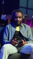 vertical jogador em sofá usando controlador e fones de ouvido para jogar videogame às lar, fechar acima. homem dentro casa teatro usando Alto tecnologia jogos console controle para derrota oponentes dentro jogo, Câmera b video