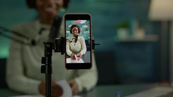 Close up of smartphone recording vlog of african influencer in home studio using smartphone. Speaking during livestreaming, blogger discussing in podcast wearing headphones and professional microphone video