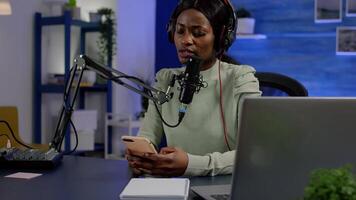 African vlogger recording blog reading messages from smartphone with modern equipment in studio podcast. Influencer looking at phone talking in broadcast using tech to connect with audiences video