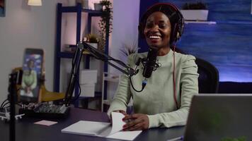 African creator content waving at camera smiling and talking with audience during podcast using smartphone. Influencer looking at phone while speaking at podcast microphone sitting in home studio video