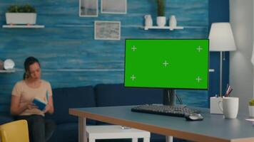 Powerful computer with mock up green screen chroma key stands on office desk in living room. Woman reading book in background in bright room with Pc isolated display video
