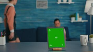 Isolated tablet computer with mock up green screen chroma key standing on desk table in living room. Two friends sitting on sofa in background talking about communication video