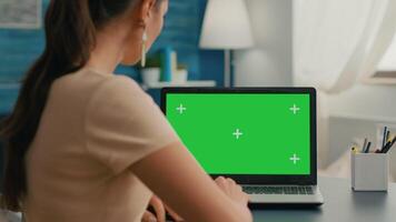Back shot of freelencer woman looking at laptop with mock up green screen chroma key while working at commerce project. Caucasian female standing in home office studio with her collegue in background video