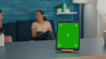 Two women sitting on sofa talking about digital technology. Office room is equipped with isolated tablet computer with mock up green screen chroma key display standing on table desk video