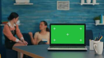 Two women sitting together on sofa in background talking about internet technology. In front on desk table standing isolated laptop computer with mock up green screen chroma key in office home video