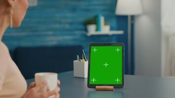 Woman drinking a cup of coffee while looking at tablet computer with mock up green screen chroma key sitting on office table. Freelancer using with isolated gadget for internet communication project video