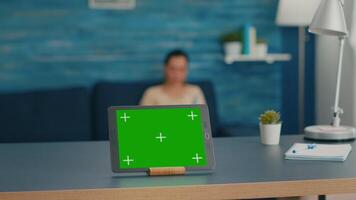 Isolated digital tablet with mock up green screen chroma key display standing on office desk in living room. In background room collegues talking about online communication video