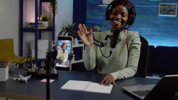 Smiling influencer african woman sitting in front of phone camera recording for blog. Digital blogger vlogger streaming talk show in studio using headphones, professional podcast microphone video