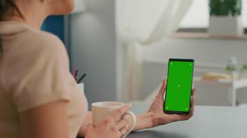 Caucasian female having online videocall using mock up green screen chroma key phone. Business woman working at online app using isolated device siiting on office desk in home office video