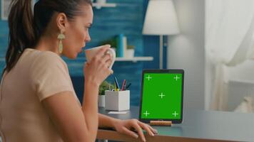 Freelancer woman holding cup of coffee looking at tablet computer with mock up green screen chroma key sitting on desk table. Caucasian female browsing on isolated device from home office room video