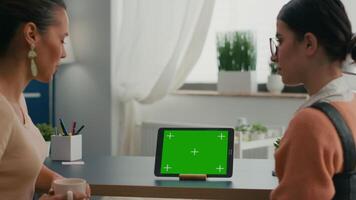 Two collegues talking about internet connection in front of tablet computer with mock up green screen chroma key. Women browsing on isolated device sittings at office desk in modern living room video