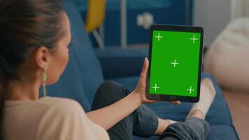 Caucasian female lying on sofa having online meeting on tablet computer with mock up green screen chroma key display. Woman using isolated touchscreen device for social networks browsing video