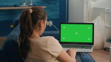 Freelancer woman sitting on sofa in home office typing on laptop computer with mock up green screen chroma key display. Caucasian woman working on financial graphs using isolated pc video