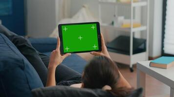 Woman sitting on sofa in living room watching movies on tablet computer with mock up green screen chroma key display. Freelancer using isolated touchscreen device for social networks browsing video