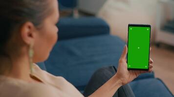 Caucasian female sitting on sofa in living room while socializes on internet app using phone with mock up green screen chroma key display.Woman browsing on network with isolated gadget video