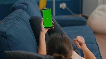 Freelancer lying on sofa while having online videocall on smartphone with mock up green screen chroma key display in vertical mode. Woman using isolated touchscreen device for social networks browsing video