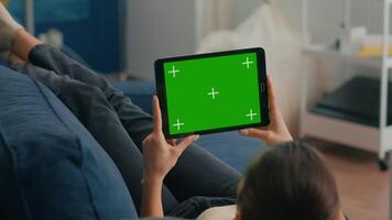Close up of woman looking at tablet computer in horizontal mode with mock up green screen chroma key display lying on couch. Freelancer using isolated touchscreen device for social networks browsing video