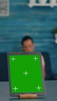 Vertical . Isolated tablet computer with mock up green screen chroma key standing on desk table in living room. Two friends sitting on sofa in background talking about communication video