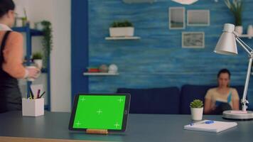 Tablet computer with mock up green screen chroma key display standing in front on office desk. Two collegues talking in background, sitting on sofa about internet connection video