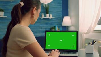 Woman typing on computer laptop with mock up green screen chroma key display working on commerce project. Freelancer using isolated pc sitting in living room while her collegue reading in background video