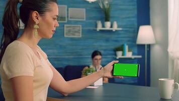 Social media person holding phone with mock up green screen chroma key display while watching online videos. Caucasian female sitting at office desk in living room using isolated technology device video