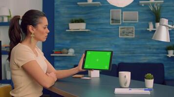 Woman watching online videos on tablet computer with mock up green screen display while sitting at office desk. Freelancer looking at template chroma key isolated device using technology internet