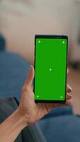 Vertical . Caucasian female sitting on sofa in living room while socializes on internet app using phone with mock up green screen chroma key display.Woman browsing on network with isolated gadget video
