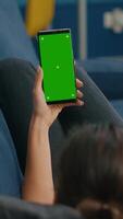 Vertical . Freelancer lying on sofa while having online videocall on smartphone with mock up green screen chroma key display in vertical mode. Woman using isolated touchscreen device for social networks browsing video
