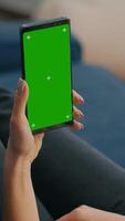Vertical . Freelancer holding in vertical mode smartphone with mock up green screen chroma key display while lying on sofa in living room. Woman using isolated touchscreen device for social networks browsing video