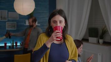 Portrait of attractive woman drinking beer during celebration wekeend. In background group of multi-ethnic friends gathering together for celebrating birthday party late at night in living room video