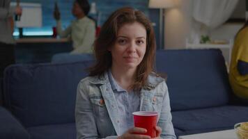 Portrait of woman sitting on sofa similing at camera while drinking beer late at night during holiday party. In background multi-ethnic friends gathering together for celebrating birthday video