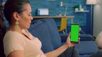 Freelancer woman sitting on sofa in living room, looking at isolated smartphone with mock up green screen chroma key in landscape mode. Caucasian female browsing on internet with friends video