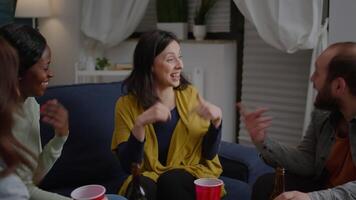 Multi-cultural friends socialising while hanging out late at night in living room during fun party. Group of mixed race people enjoying time spending together cheering beer bottles video