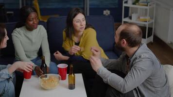 Multi-cultural friends socializing while sitting on sofa in living room late at night drinking beer enjoying home party. Group of mixed race people hanging out enjoying time spending together video