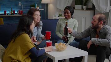 Multi-ethnic friends spending time together, drinking beer during nightlife party while sitting on sofa late at night in living room. Group of mixed race people hanging out, having fun conversation video