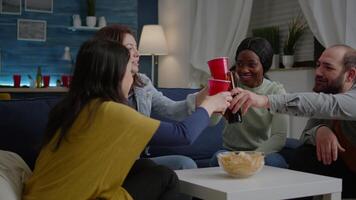 Group of multi-cultural friends having fun during entertainment wekeend party drinking beer, eating snack while sitting on sofa. Mixed race people hanging out enjoying time spending together video