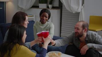 Multi-cultural friends cheering bottles of beer spending time together while relaxing on couch. Group of mixed race people hanging out, socializing during wekeend party late at night video