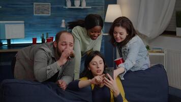 Multiracial friends hanging out late at night watching funny entertainment on smartphone while relaxing on couch in living room. Group of multiethnic people spending time together having fun video