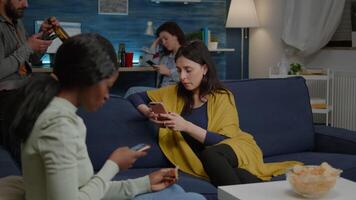 Multiracial friends relaxing on couch while surfing news on internet using mobile speding time together during surprise party. Group of multi-ethnic people watching online funny movie in living room video