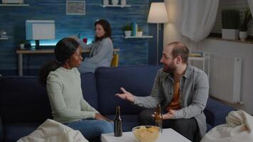 Multi-ethnic friends relaxing on couch while giving high five laughing and having fun during night celebration birthday. In background women gathering together socialising during wekeend party video