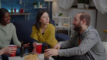 Multiracial friends laughing while enjoying night party drinking beer, sharing funny story relaxing on sofa late in living room. Group of mixed race people hanging out spending time together video
