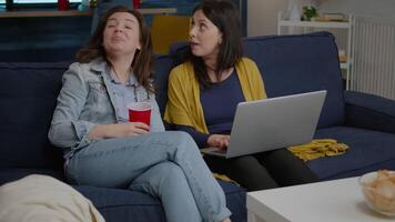 Group of multiethnic people discussing while watching entertainment on laptop resting on couch in living room. In background afro american woman and man drinking beer enjoying free time together video