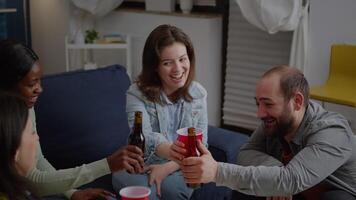 Multi cultural friends cheering beer bottles during wekeend party laughing while relaxing on sofa late at night. Group of mixed race people hanging out enjoying time spending together video