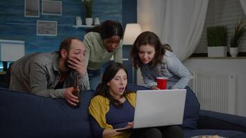 Multi-ethnic friends watching interesting comedy movie on laptop computer relaxing on couch, drinking beer, having fun. Group of mixed race people spending time together during night party video