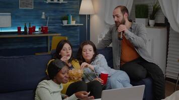 Multiracial friends chilling on sofa in living room during movie night watching online comedy film using laptop computer. Group of mixed people hanging out together, drinking beer, eating snacks video
