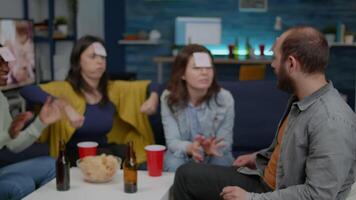 Mixed race friends socializing during creative party while playing funny guess who game using sticky notes. Mixed race people having fun, laughing together while sitting on sofa late at night video