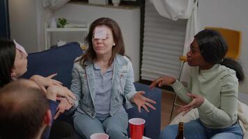 Multi ethnic friends drinking beer while playing guess who game using sticky notes. Group of multiracial people spending time together having fun during entertainment wekeend video